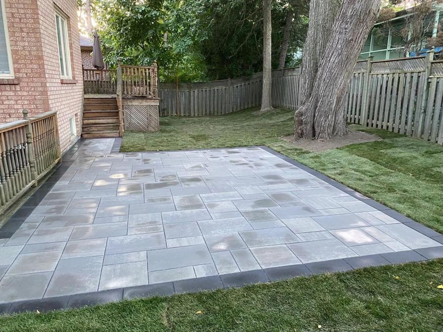 Grey paver patio with a charcoal border beside a brick house and lawn
