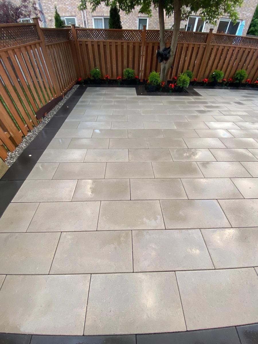 Grey paver patio with a charcoal border and a planting bed around a tree along a wooden fence