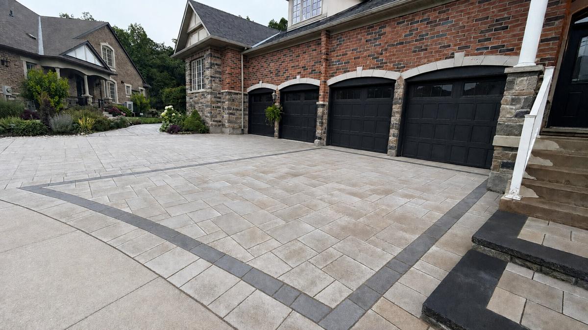 Wide interlock driveway with a sweeping charcoal border in front of a brick home’s garage bays