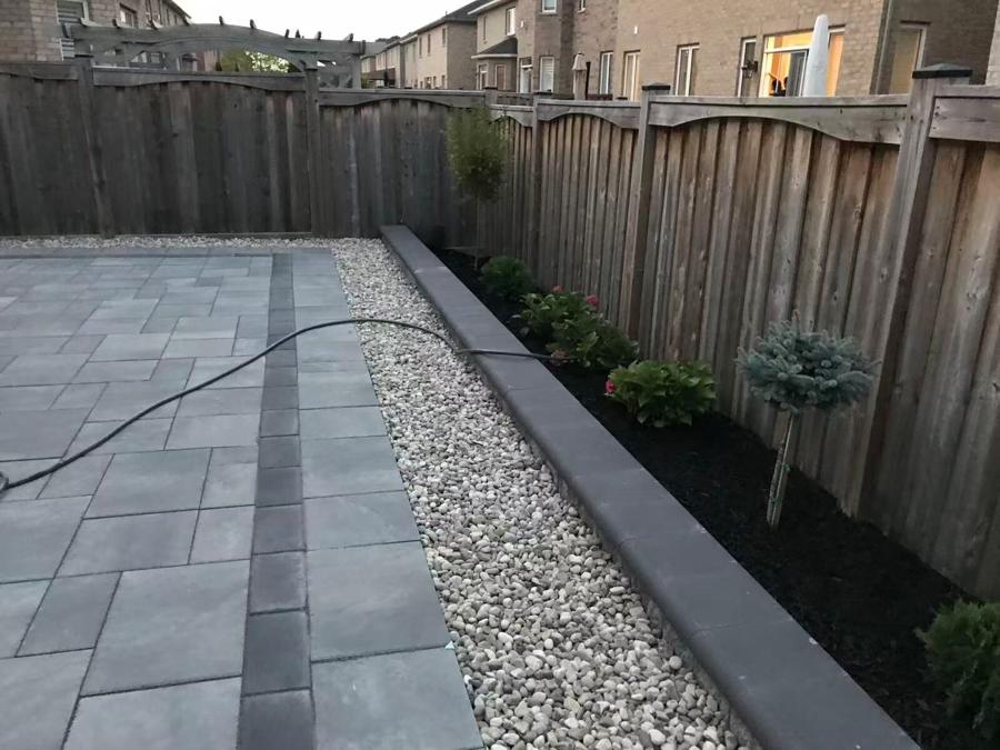 Paver patio edged with a white river-rock strip and a planted bed along a cedar fence