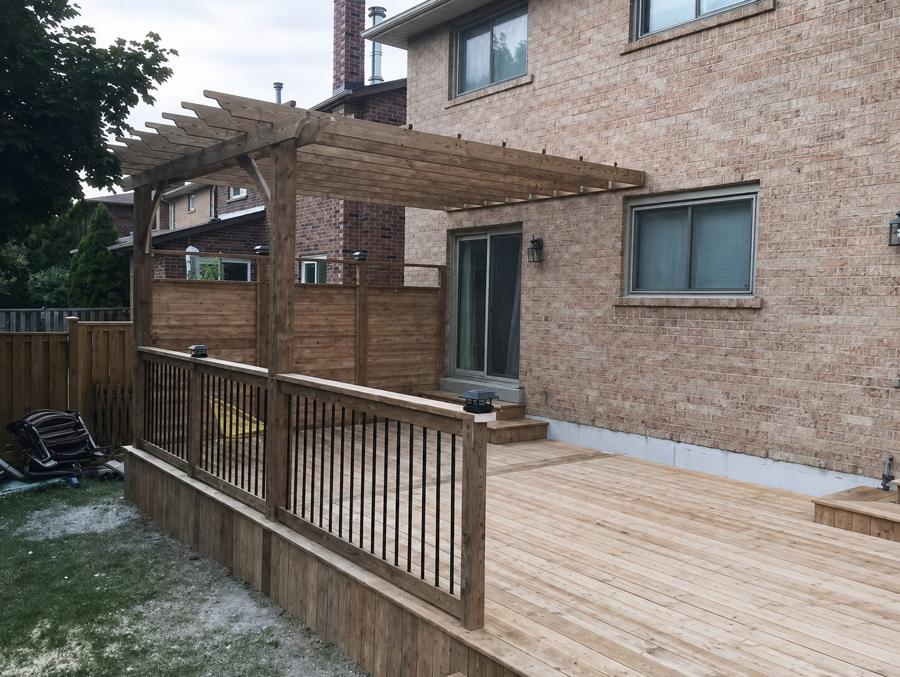 Wood pergola over a deck with black metal balusters beside a brick house