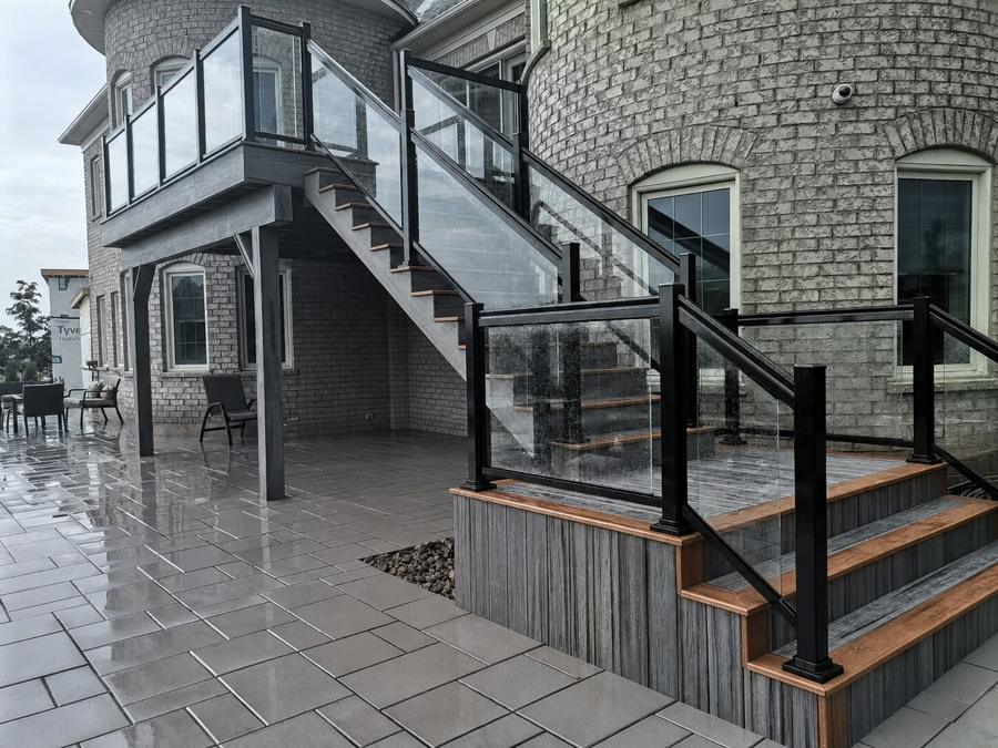 Multilevel grey deck and staircase with black-framed glass railings above a paver patio