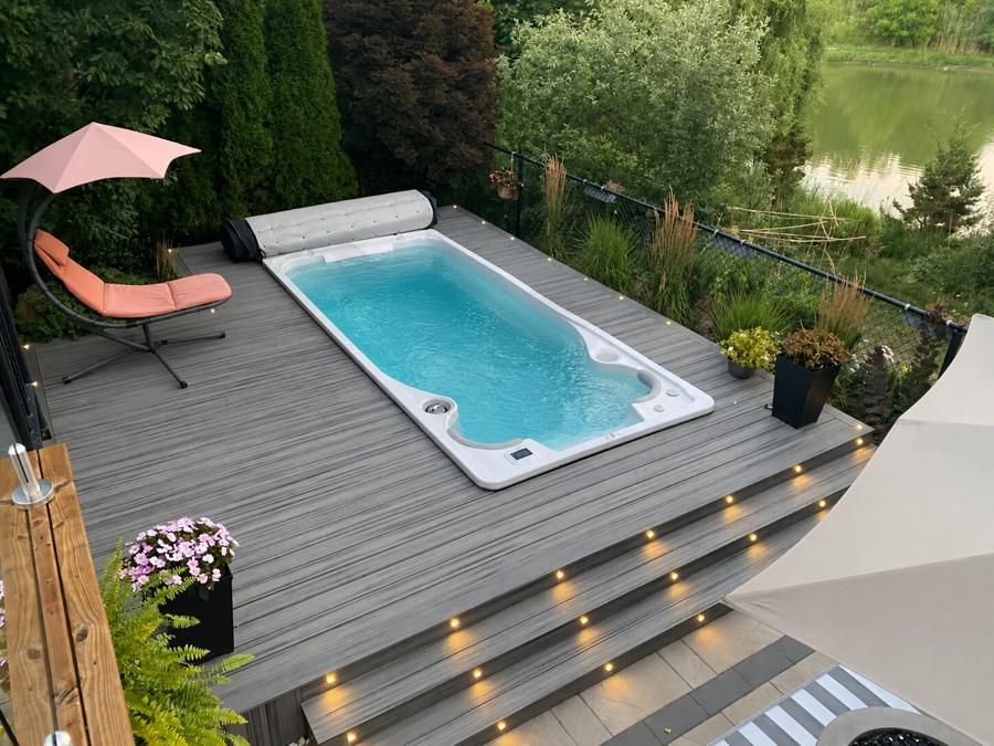 Swim spa set into a grey deck with illuminated steps, flower planters and a view toward a pond