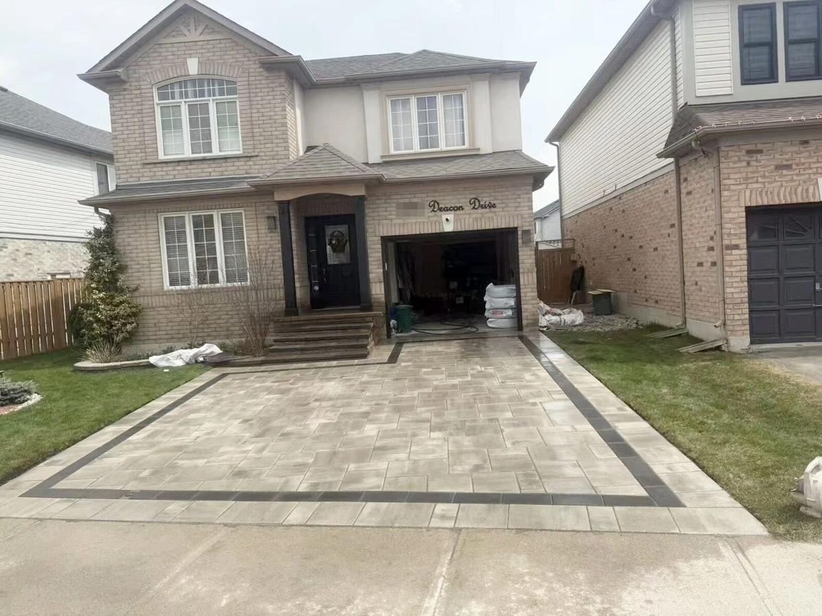 A finished residential interlock driveway in light beige pavers with a charcoal border, leading to a two-storey brick home's garage.