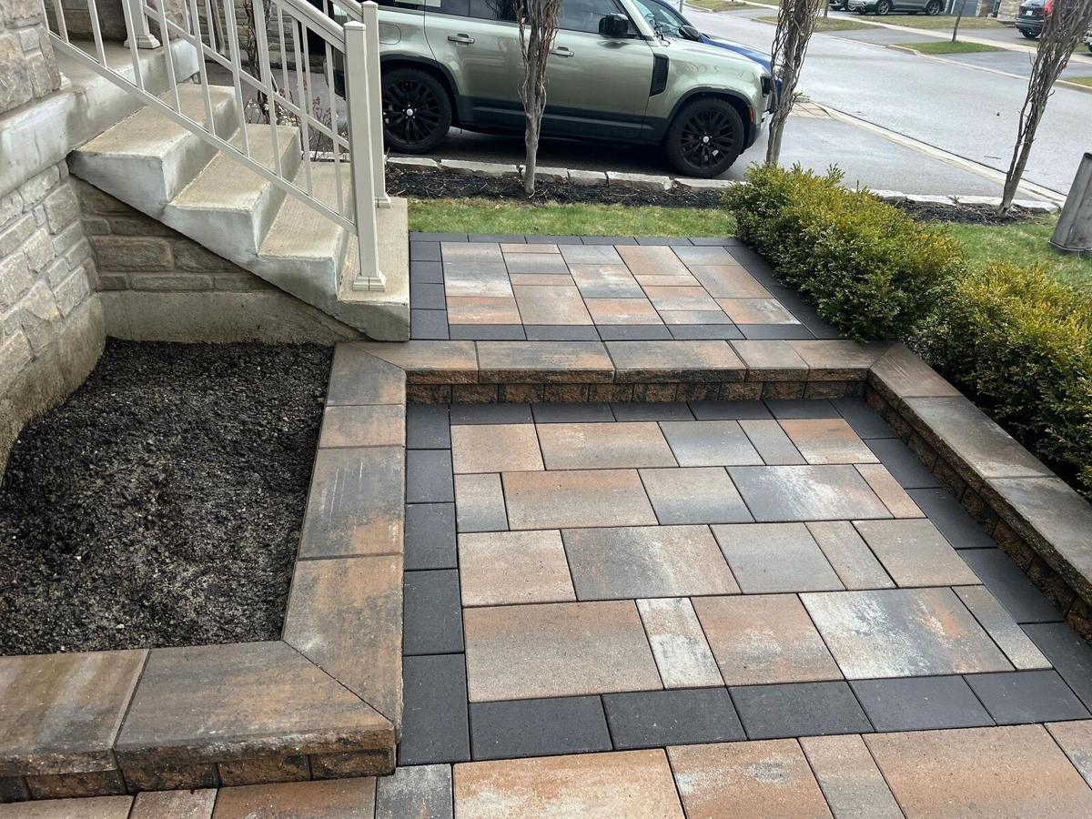 A finished front-entrance interlock landing in warm multi-tone pavers with a charcoal border, beside stone steps and a mulched garden bed.