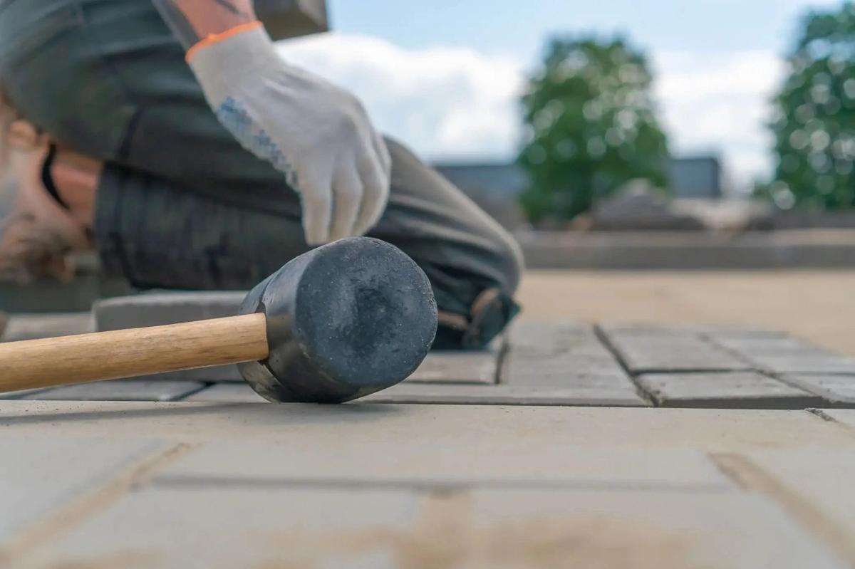 Close-up of a rubber mallet tapping interlock pavers into place, a gloved installer kneeling behind.